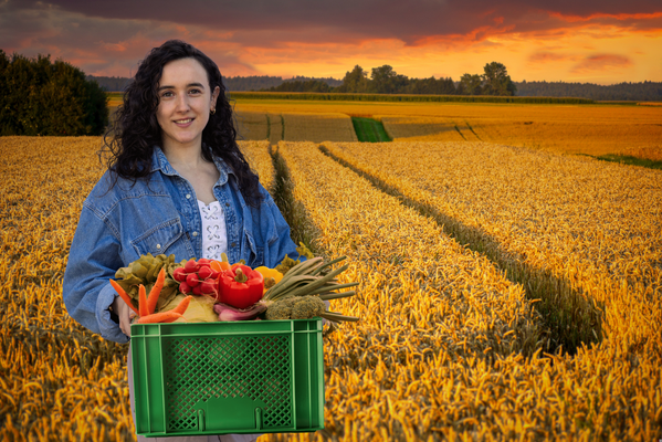 KI generiert: Frau hält eine Kiste mit frischem Gemüse auf einem Getreidefeld bei Sonnenuntergang.